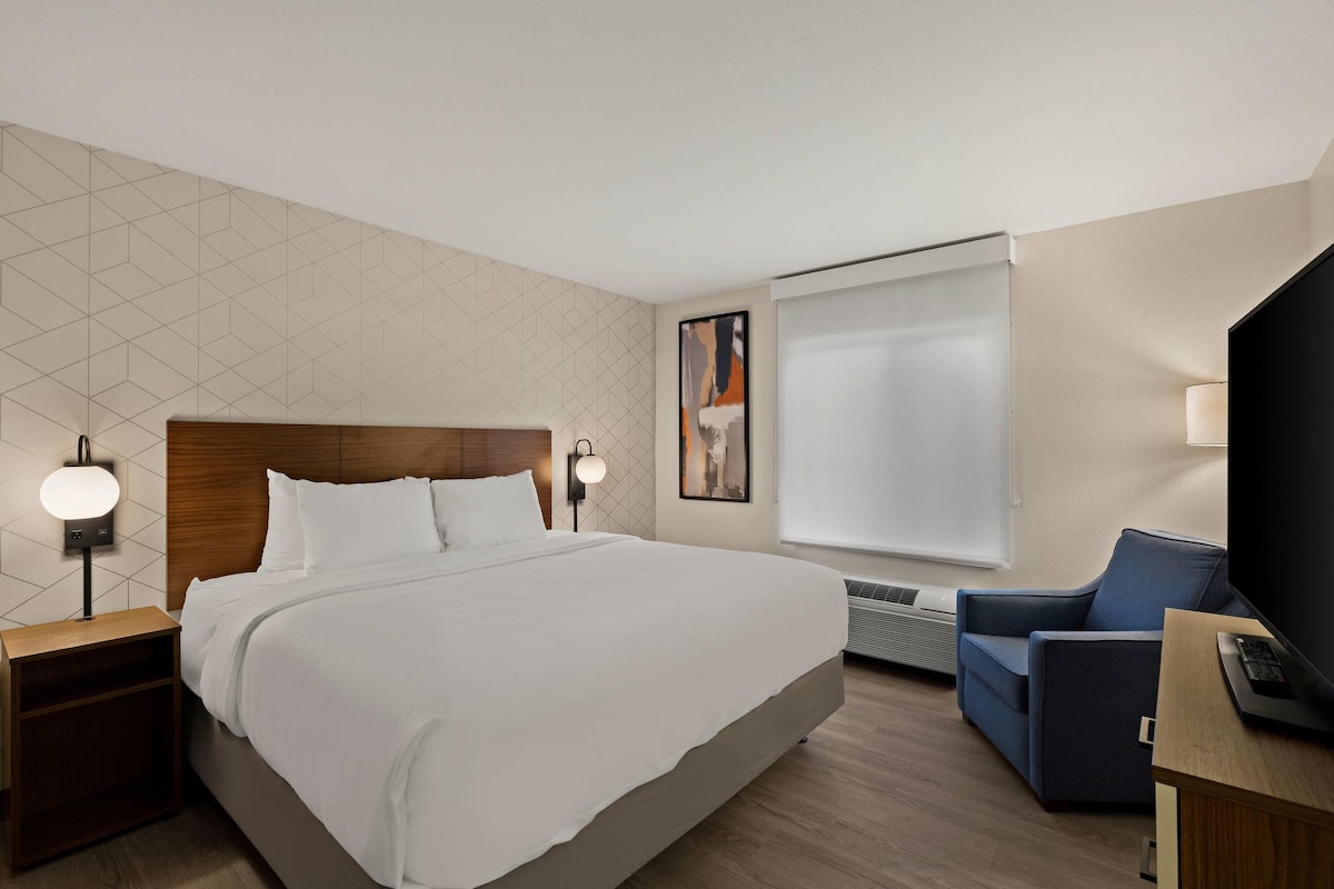 A hotel room with a large bed, a desk with a computer, a chair, a TV, and a window with blinds.