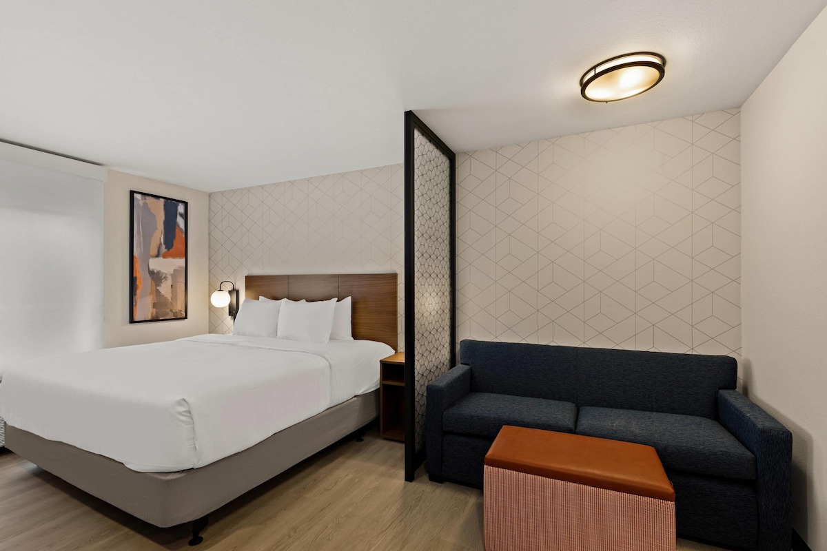 A hotel room with a bed, a sofa, a small table, a nightstand, and a wall-mounted light fixture.