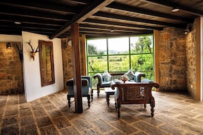 A rustic room with stone walls, a wooden ceiling, a large window with a view of greenery, and traditional furniture.