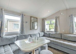 Ein Zimmer mit einer grauen Eckcouch, einem kleinen Tisch mit einer Kanne und Tassen sowie einem Fenster mit Vorhängen.
