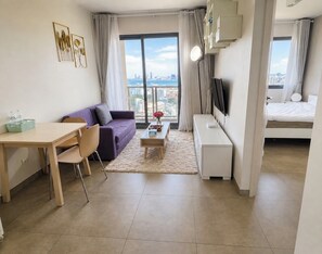 A modern hotel room with a dining area, a sofa, a TV, and a balcony with a city view. - 1 BR Sea View Condo Near Walking Street (ตำบล หนองปรือ)