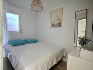 Ein Schlafzimmer mit einem Bett, einem Fenster, einem Wandbild, einem Spiegel und einem Nachttisch mit einer Vase.