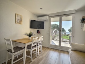 Ein Essbereich mit Holztisch und weißen Stühlen, einem an der Wand befestigten Flachbildfernseher und einer Glastür zu einem Außenbalkon.