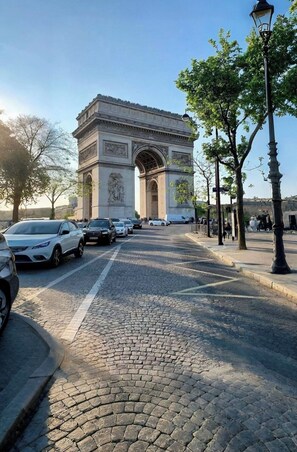 Une rue pavée menant à une grande arche historique, avec des voitures garées le long du trottoir.