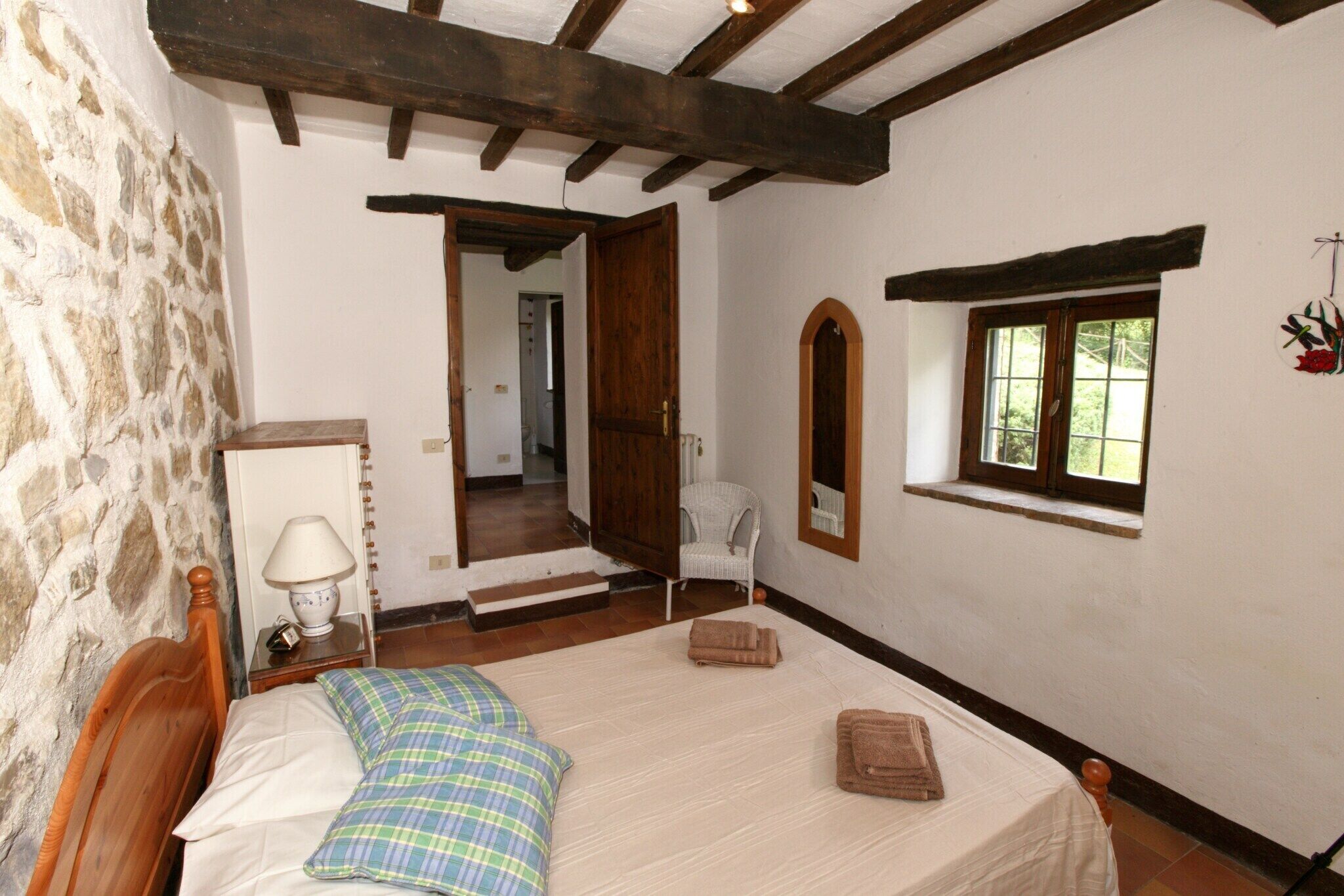 Un dormitorio con cama de madera, mesita de noche, lámpara, silla, ventana y pared de piedra.