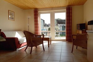 Ein Zimmer mit einer Couch, zwei Korbstühlen, einem kleinen Tisch, einem Fernseher und Blick durch eine Glastür.