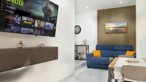 Een moderne woonkamer met een flatscreen televisie waarop een film draait, een blauwe bank met oranje kussens, een houten dressoir en een eettafel met stoelen.