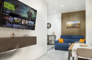 A modern living room with a flat-screen TV displaying a movie, a blue sofa with orange cushions, a wooden sideboard, and a dining table with chairs. - BorgoNaro, an authentic oasis of peace located in the charming village of Dattilo. (Dattilo)