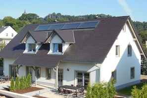 Ein zweistöckiges Haus mit geneigtem Dach, Solaranlage, einer Terrasse mit Tisch und Stühlen sowie einem kleinen Garten.