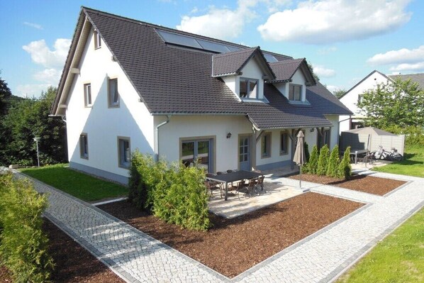 Ein zweistöckiges Haus mit gepflasterter Auffahrt, einer Terrasse mit Tisch und Stühlen sowie einem kleinen Garten.