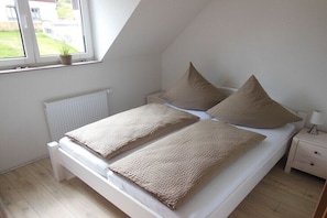 Ein Schlafzimmer mit einem Bett, Kissen, einem Nachttisch, einer Lampe, einem Fenster mit Ausblick und einer Heizung.