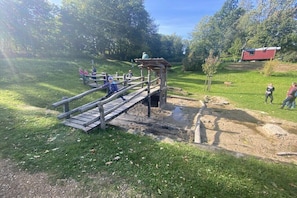 Eine Holzbrücke über einem Bach, umgeben von Grün und ein rotes Gebäude im Hintergrund.