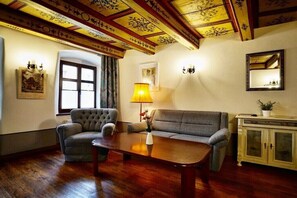 Ein Zimmer mit Holzbalkendecke, einer Couch, einem Sessel, einem Tisch und einem Schrank.