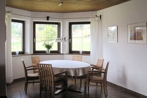 A dining area with a round table, six chairs, and three windows with curtains. - Heidi Pfeifhofer - Apt. B (Bodensdorf am Ossiacher See)