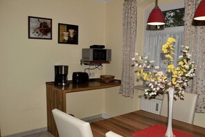Uma sala de jantar com uma mesa de madeira, cadeiras, uma cafeteira, micro-ondas e um vaso com flores.