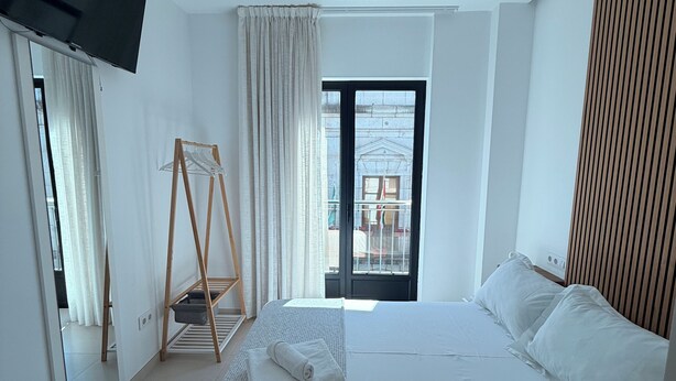 A modern bedroom with a large bed, a TV mounted on the wall, a balcony with a view, and a wooden slat wall.