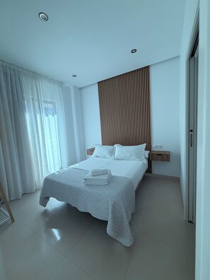 A modern hotel room with a neatly made bed, a wooden headboard, a nightstand, and a large window with sheer curtains.