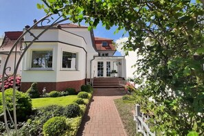 Ein zweistöckiges Gebäude mit weißer Fassade und rotem Dach, ein Hauseingang mit Treppe und ein Garten mit verschiedenen Pflanzen und einem Weg.