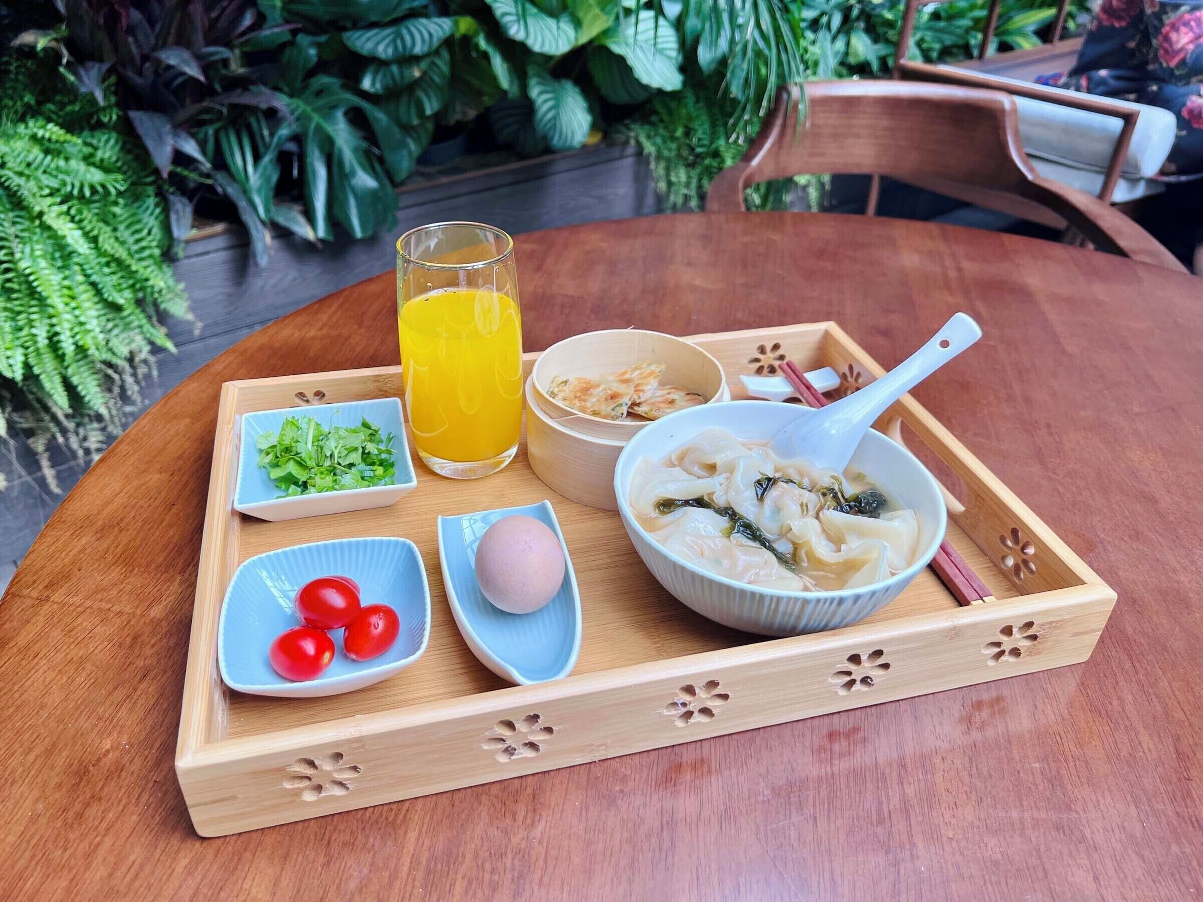 A wooden tray with a bowl of noodles, a glass of orange juice, a bowl of greens, a small bowl of cherry tomatoes, and a pink egg on a table.