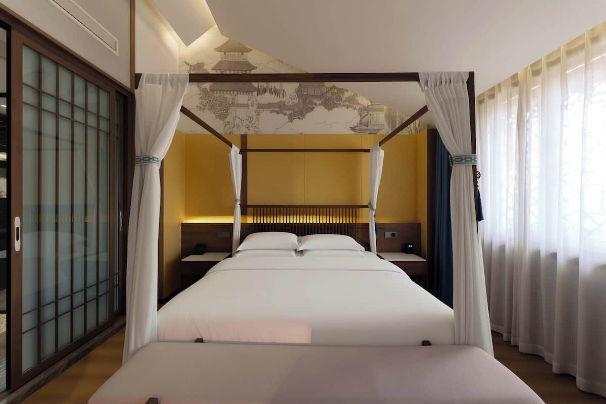 Una cama con dosel, ropa de cama blanca, cabecera de madera y vista a un edificio tradicional a través de una ventana con cortinas blancas.