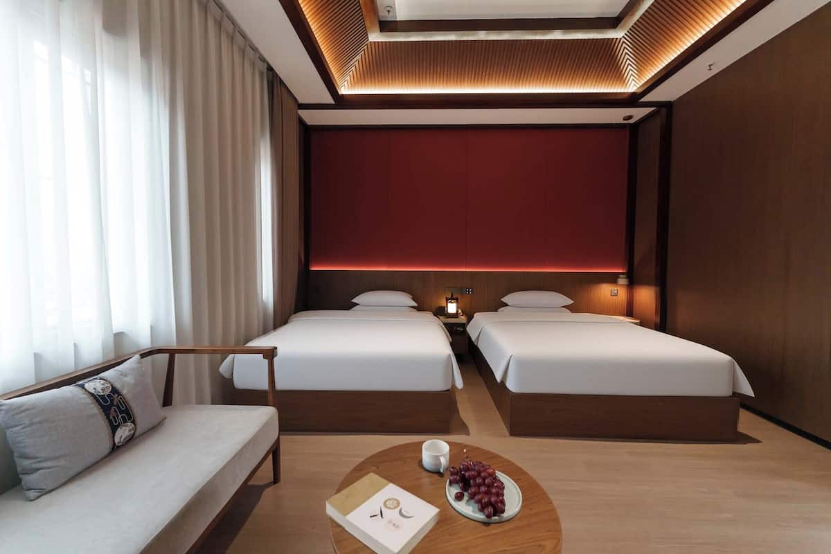 Habitación de hotel con dos camas, un sofá, una mesita de noche con lámpara y vistas a la ciudad.