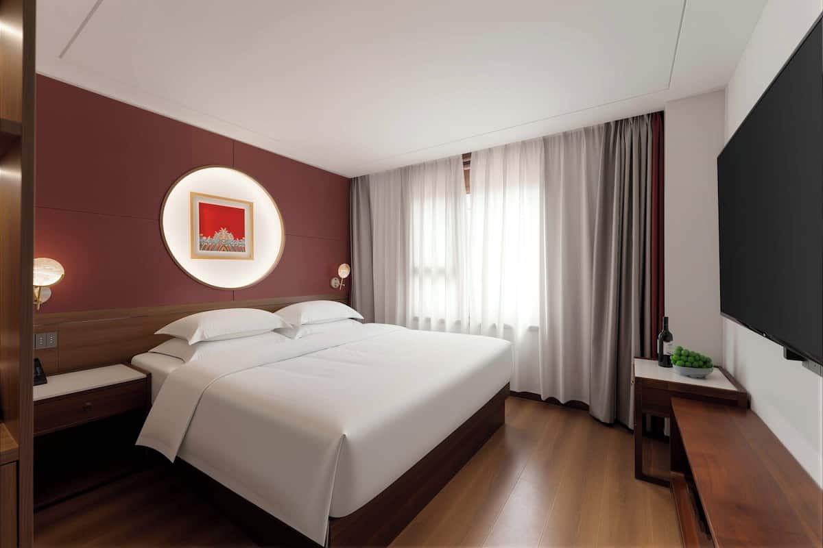Habitación de hotel con una cama grande, una mesita de noche, un televisor y una ventana con cortinas.
