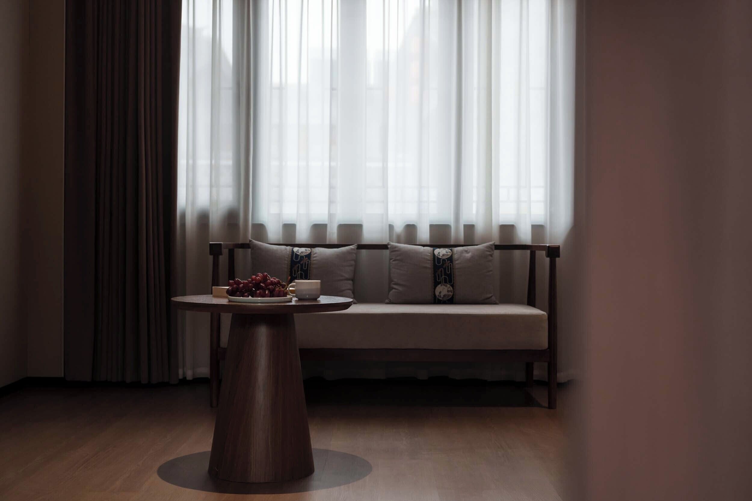 A room with a wooden floor, a round table with a white cup and a bowl of fruit, and a sofa with patterned cushions.