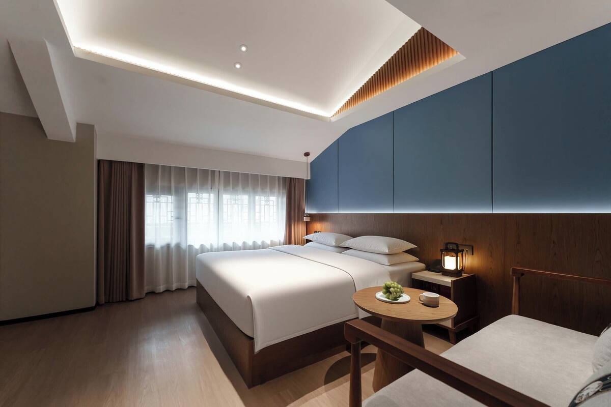 Una habitación de hotel moderna con una cama grande, mesita de noche, lámpara y una pequeña mesa redonda con una taza y fruta.