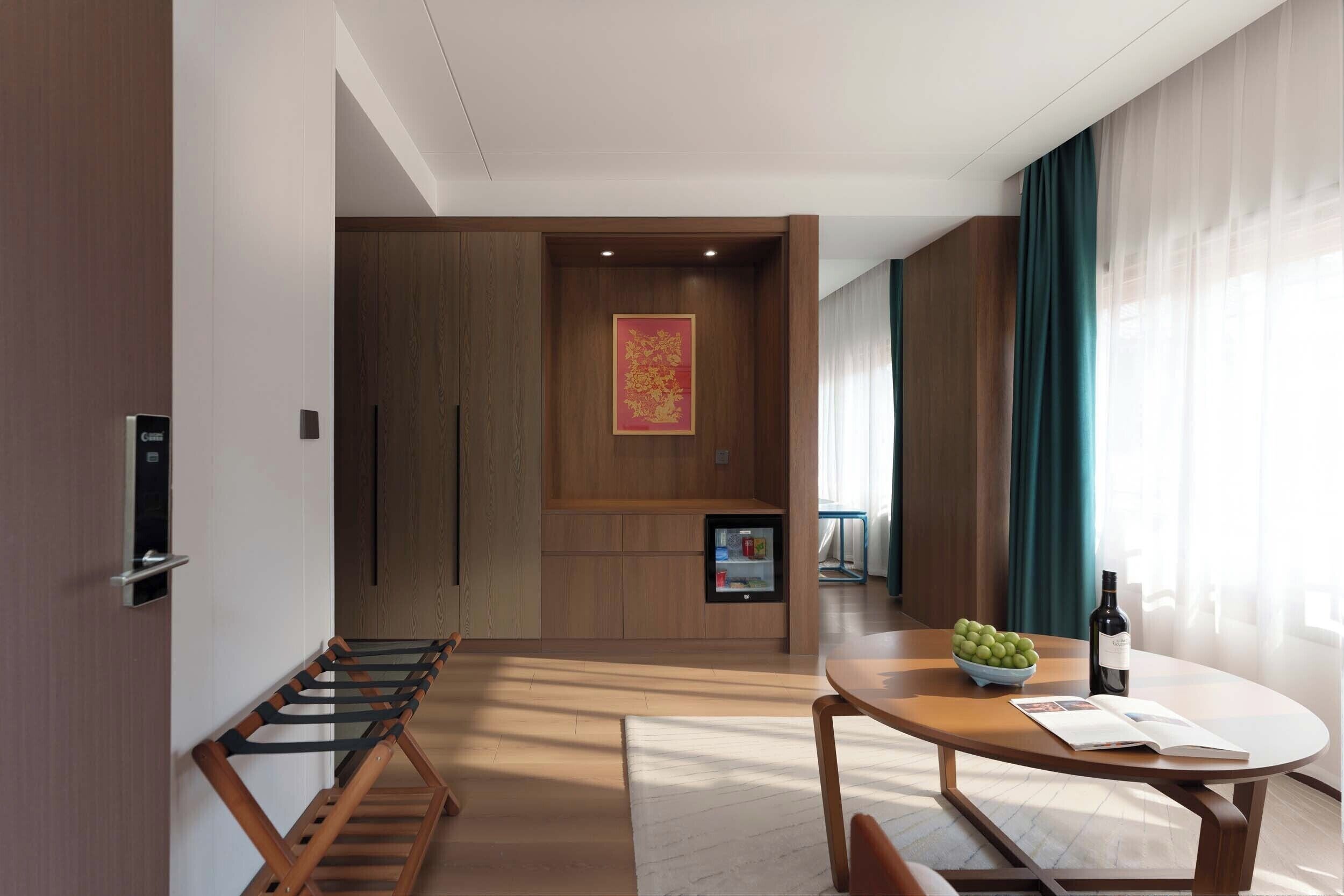 Una habitación de hotel moderna con una pared de madera, una pequeña mesa con un cuenco de fruta y una botella, un banco y una puerta con un panel de control.