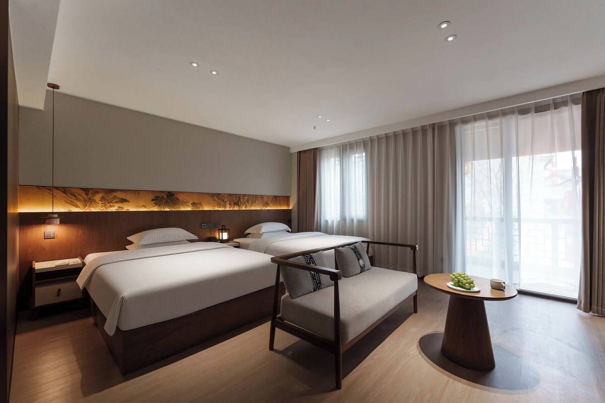 Habitación de hotel con dos camas, un sofá, una mesa redonda con frutero y un ventanal grande con cortinas.