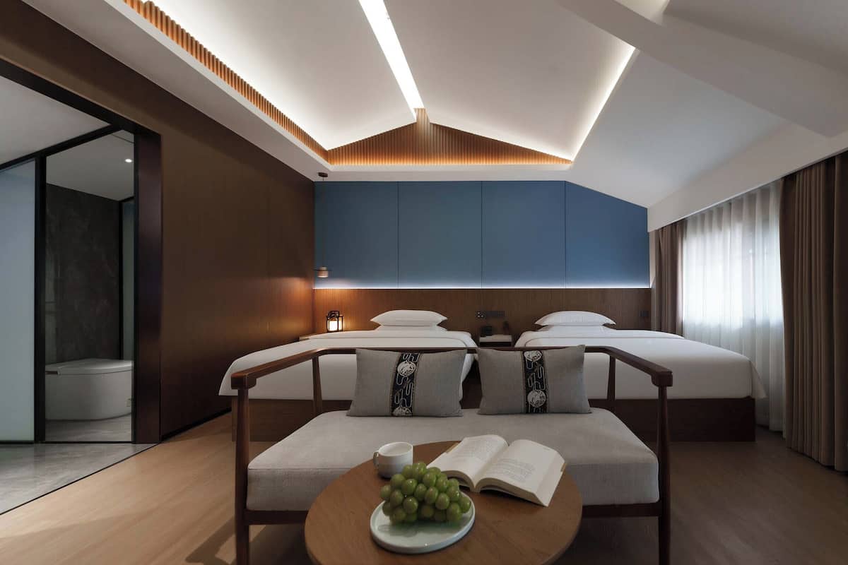 Habitación de hotel con cama, mesitas de noche, una zona de lectura con un libro y uvas, y un baño visible a través de una puerta abierta.