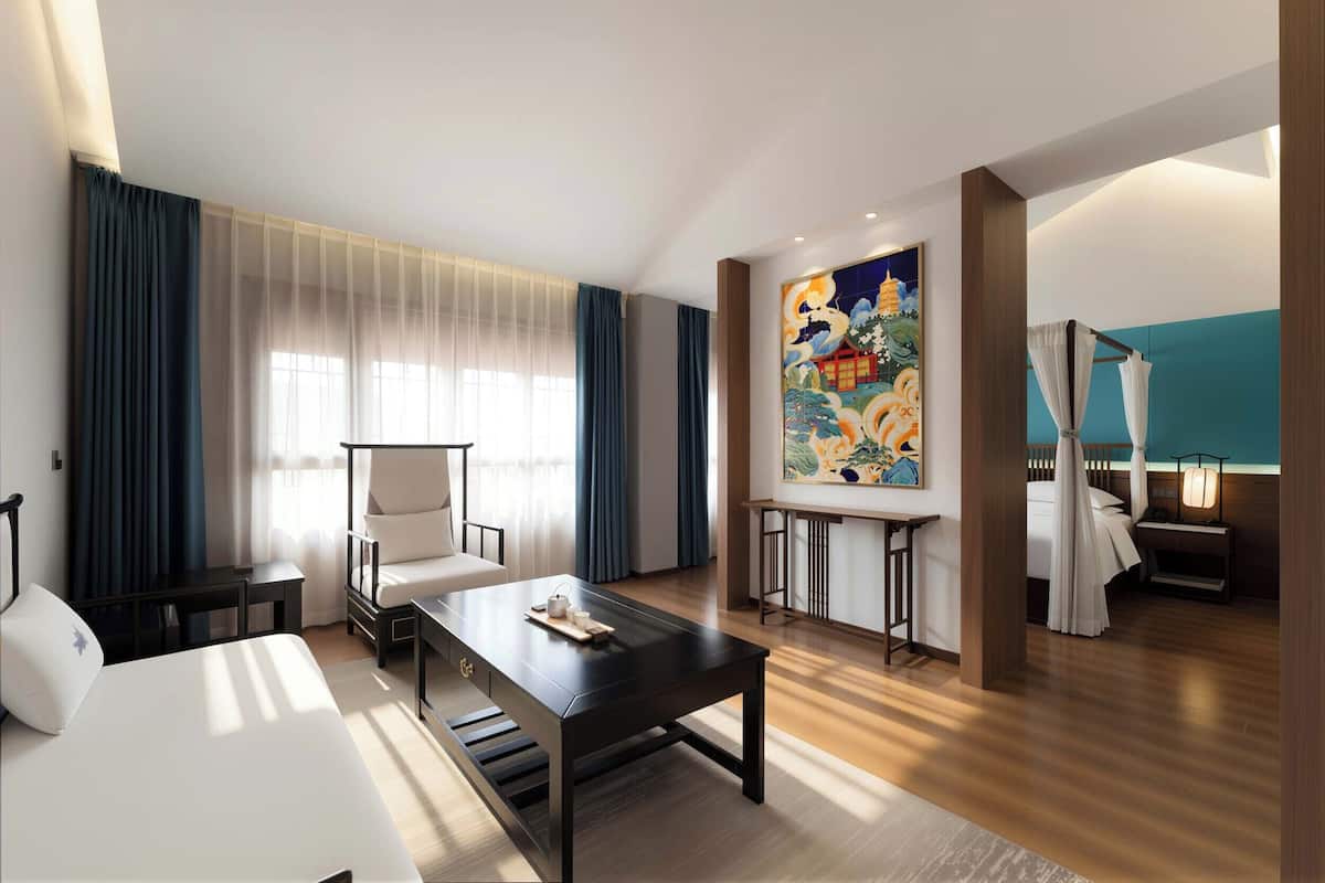Habitación de hotel con cama, escritorio, silla, un cuadro y ventana con cortinas.