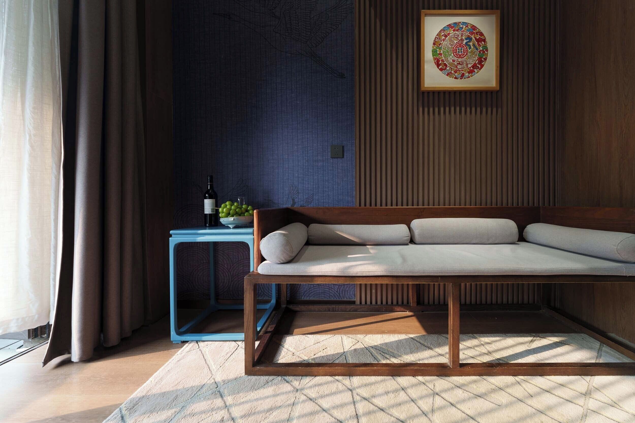 A modern living room with a sofa, a small side table with a bottle and fruit, and a wall with a decorative panel and framed artwork.