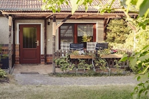 A house with a front door, a small porch, a wooden table with chairs, and a garden area with plants and flowers. - Merle in Plau - Heidenholz (Plau am See)