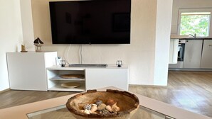 Una sala de estar moderna con un televisor de pantalla plana, un cuenco de madera con objetos decorativos, un mueble bajo de color blanco y una zona de cocina con armarios.