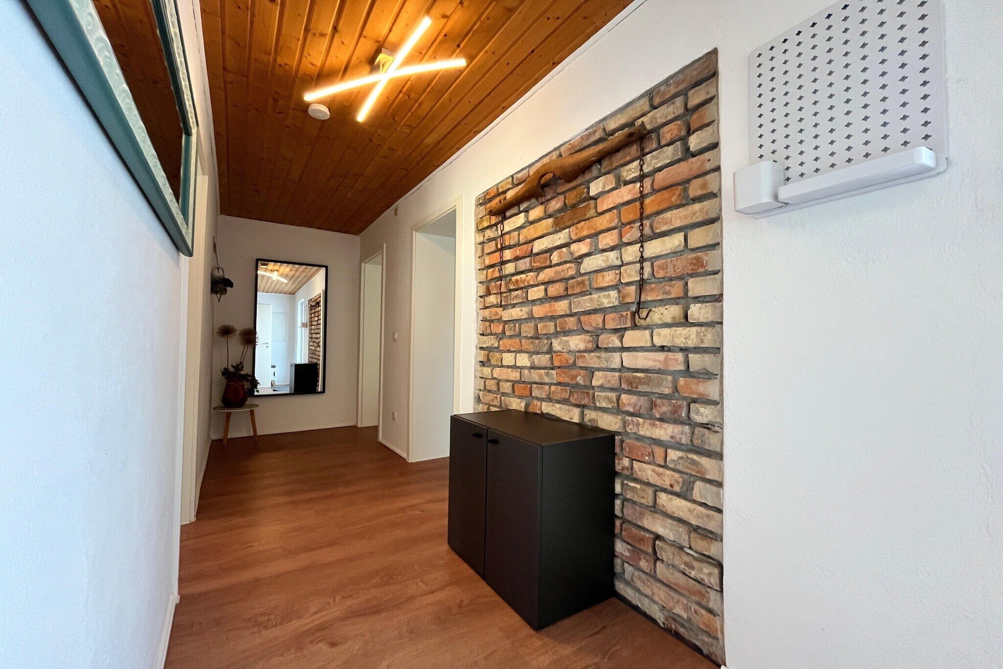 A hallway with wooden flooring, a brick accent wall, a black cabinet, and a ceiling with exposed beams and lighting.