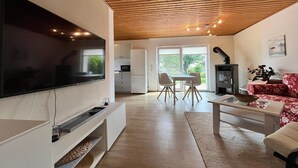 Una sala de estar moderna con un televisor de pantalla plana, un comedor con mesa y sillas, una cocina al fondo y un techo de madera.
