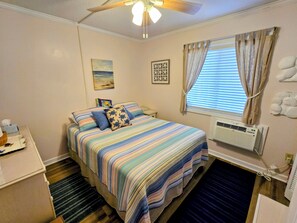 Ein Schlafzimmer mit einem Bett, einem Deckenventilator, einem Fenster mit Vorhängen, einer Klimaanlage und einem kleinen Schreibtisch mit Lampe.