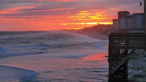 Tramonto sulla costa con onde, un molo di legno e edifici sullo sfondo.