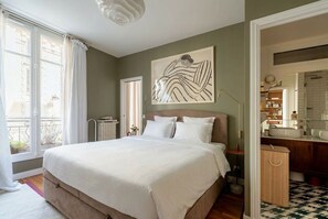 Ein Schlafzimmer mit Bett, einem großen Fenster, einem Bild an der Wand und einem durchgehenden Tür angedeuteten Badezimmer.
