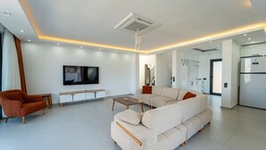 Ein modernes Wohnzimmer mit einem Ecksofa, einem Fernseher, einem Couchtisch und einem Essbereich mit einem Kühlschrank.