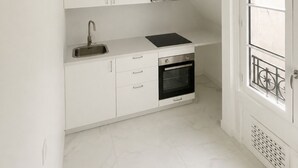A compact kitchen with white cabinetry, a built-in oven, and a window with a metal railing. - COSY, FULLY-EQUIPPED APARTMENT (Paris)