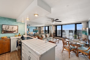 A spacious living area with a large window, a dining table with chairs, a kitchen with a tiled island, and a ceiling fan. - West Maui 1BR condo with pool, AC, kitchen, and oceanfront access-Kaleialoha 306 (Lahaina)