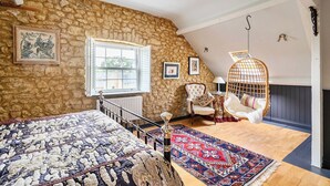 Un dormitorio con pared de piedra, una cama con colcha estampada, una silla colgante, una butaca con cojín, una ventana y una lámpara colgante.