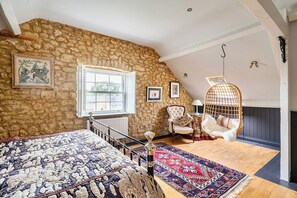 Un dormitorio con pared de piedra, una cama con colcha estampada, una silla colgante, una butaca con cojín, una ventana y una lámpara colgante.