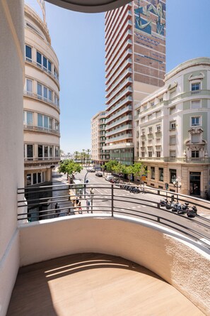 Blick von einem Balkon auf eine Stadtstraße mit Gebäuden, einem klaren Himmel und wenigen Fußgängern.