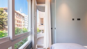 Ein Schlafzimmer mit großem Fenster, einem Bett und Blick auf Gebäude und Grünflächen.