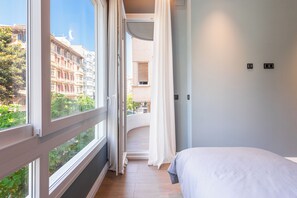 Ein Schlafzimmer mit großem Fenster, einem Bett und Blick auf Gebäude und Grünflächen.