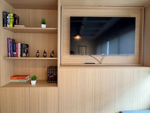 Ein modernes Wohnzimmer mit einem an der Wand befestigten Fernseher, Holzregalen mit Büchern und Dekoration sowie einem kleinen Topfpflanzen.
