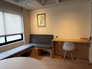 Ein Zimmer mit einer Bank, einem Schreibtisch, einem Stuhl, einer kleinen Pflanze und einem gerahmten Kunstwerk an der Wand.